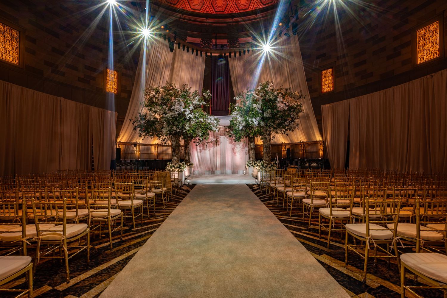 Wedding ceremony venue with rows of wooden chairs and floral arrangements leading to an illuminated altar area.