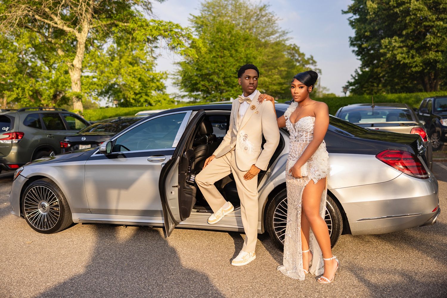 A couple poses elegantly next to a luxury silver Mercedes-Benz sedan at an outdoor event.