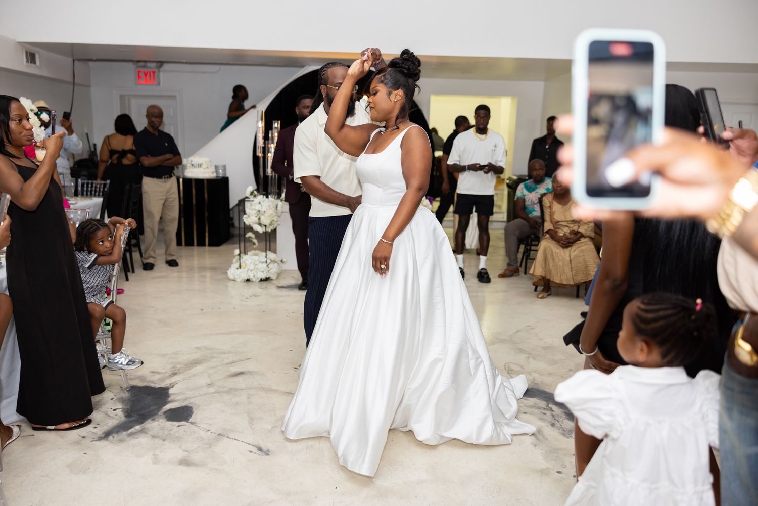 A person in a flowing white wedding dress dances joyfully on the dance floor while guests take photos on phones.