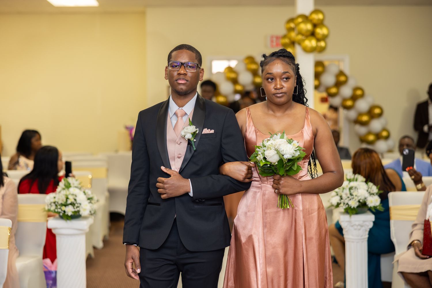Wedding party in formal attire poses in reception venue with gold balloon decorations.