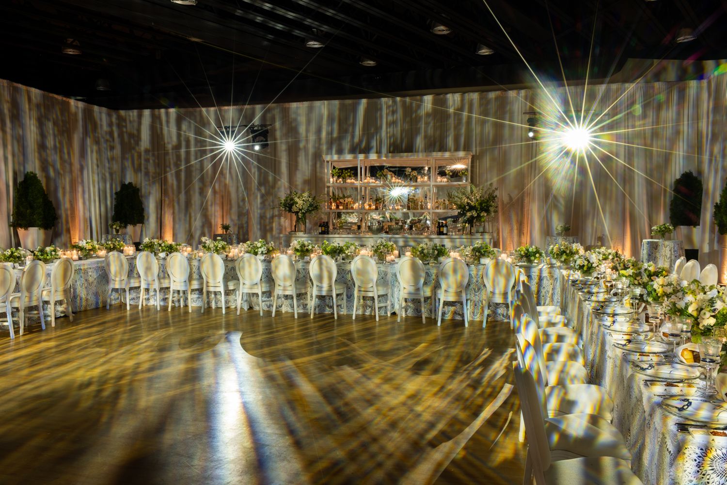 Elegant ballroom with wooden floors and dramatic lighting features crystal chandeliers and floral centerpieces.