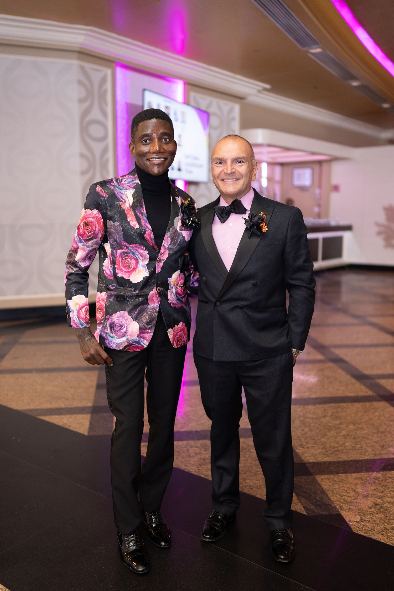 Two people pose together at an event wearing contrasting floral and classic tuxedos under pink lighting.