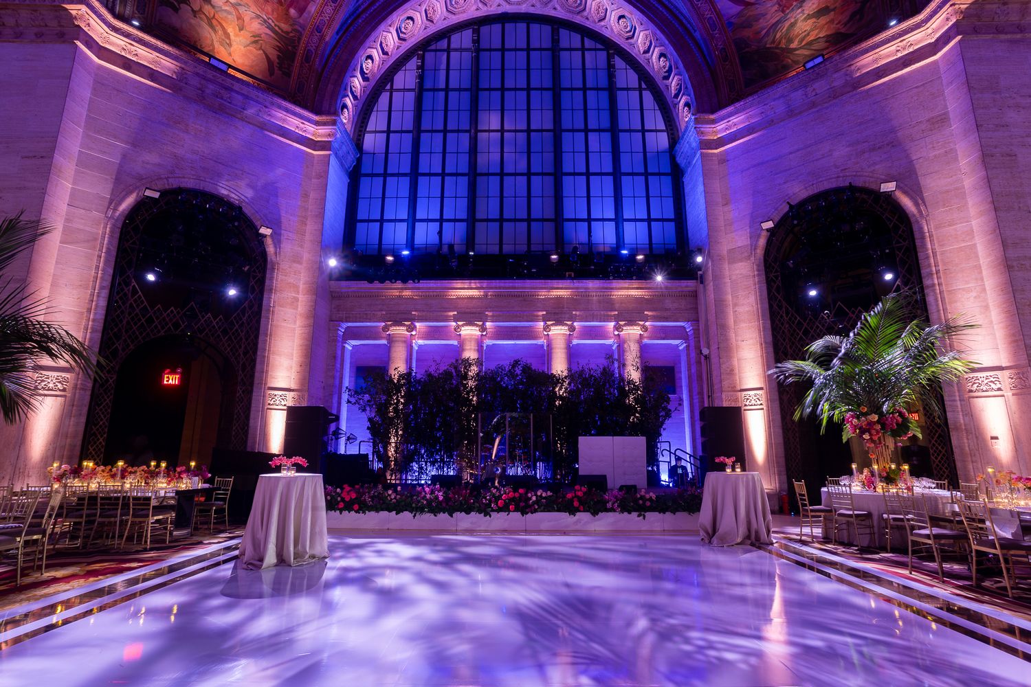 A luxurious ballroom with purple uplighting and elegant decor features dramatic floor-to-ceiling windows and columns.