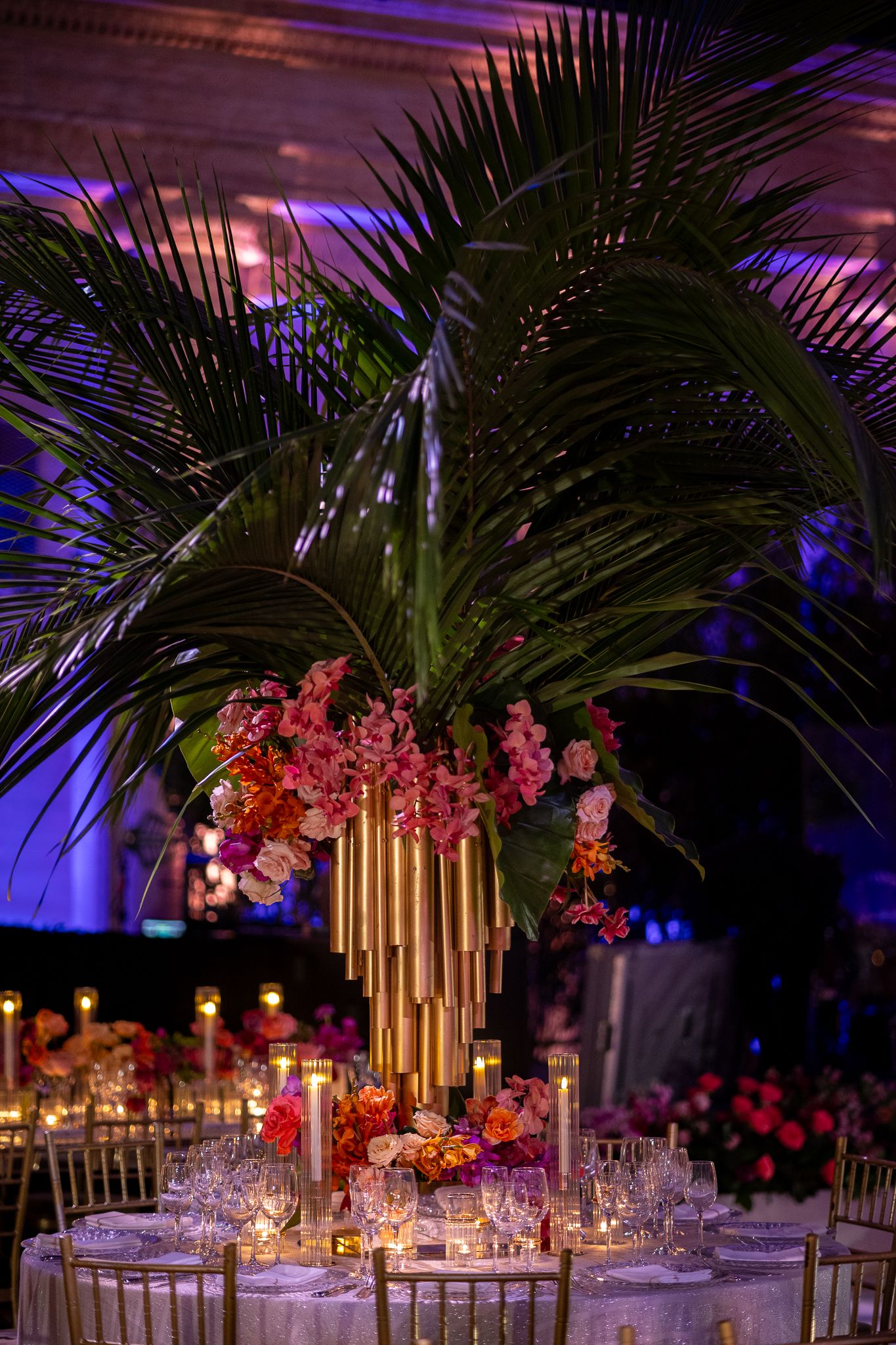 Elegant wedding reception tables decorated with tall palm fronds and pink floral arrangements under purple mood lighting.