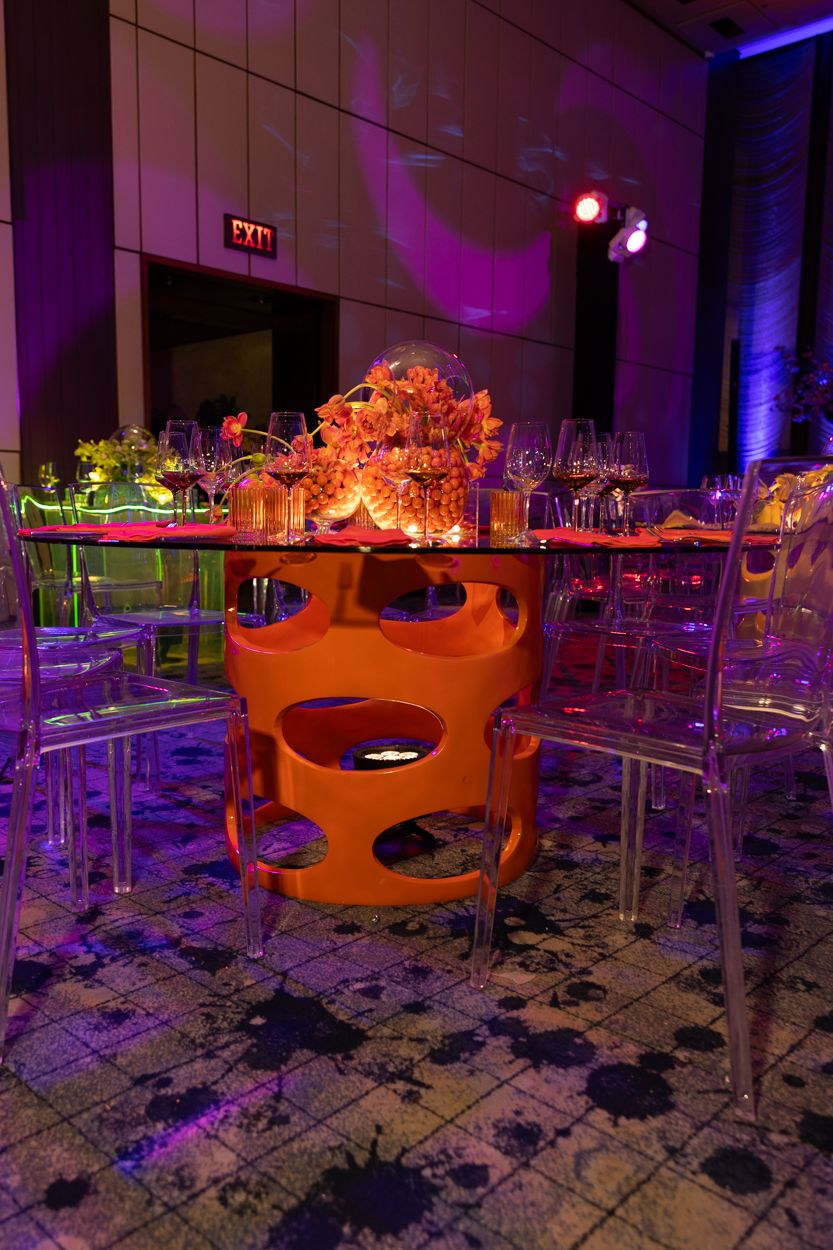Modern event space with orange floral centerpieces and purple lighting illuminating clear acrylic chairs and patterned floor.