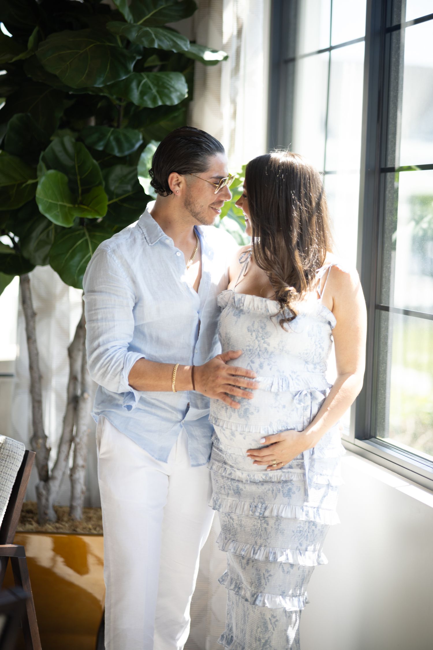 Couple in white and light blue outfits share an intimate moment by a window with indoor plants.