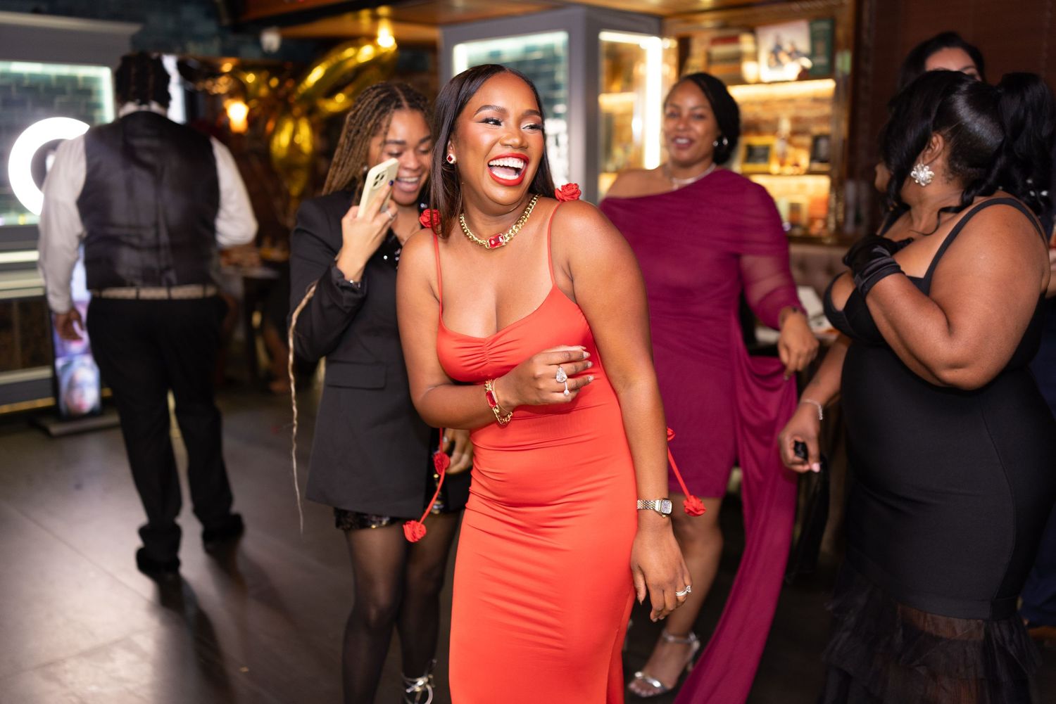A woman in a bright coral dress laughs joyfully while dancing at a lively indoor party venue.