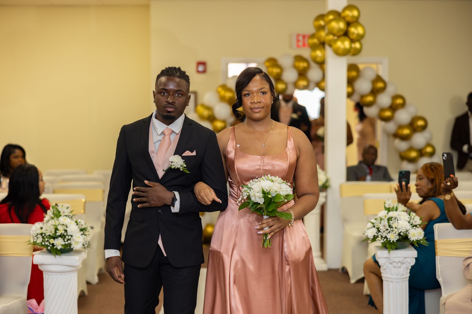 Wedding party members in pink dresses and formal suits celebrate with gold balloons in an elegant venue.