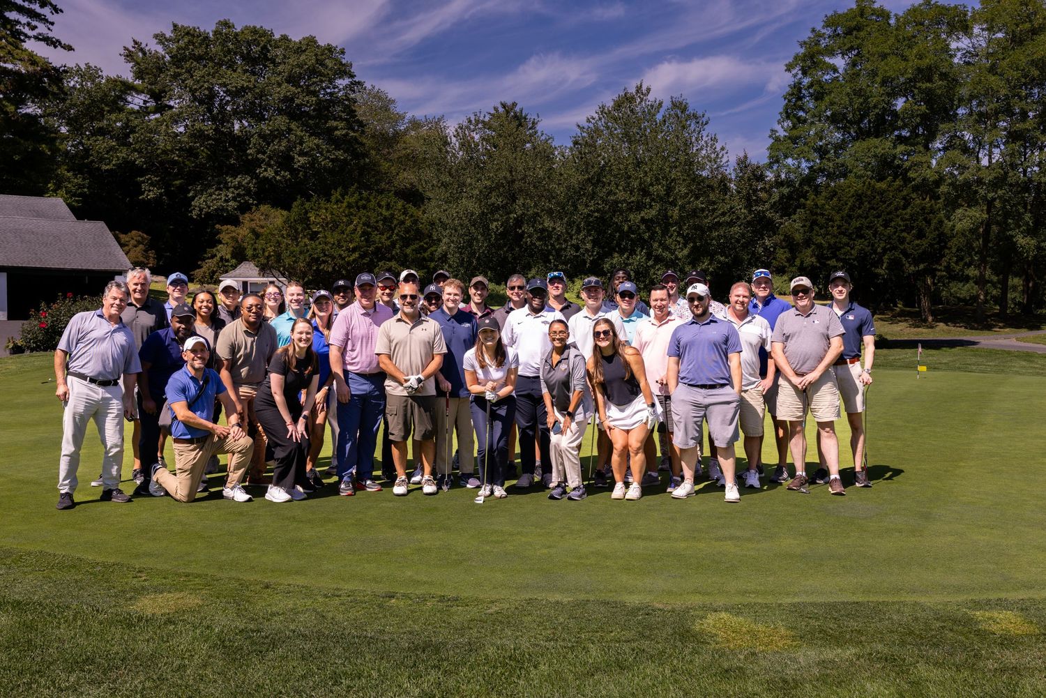 A large group of people in casual attire sitting together on a grassy lawn during a sunny day.