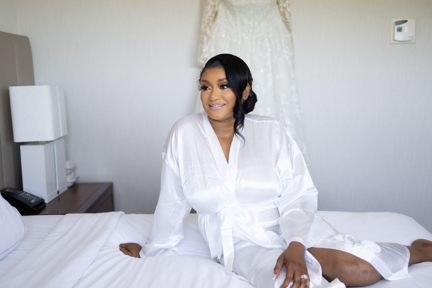 Someone wearing a white satin robe sits on a bed in a hotel room with a wedding dress visible in the background.