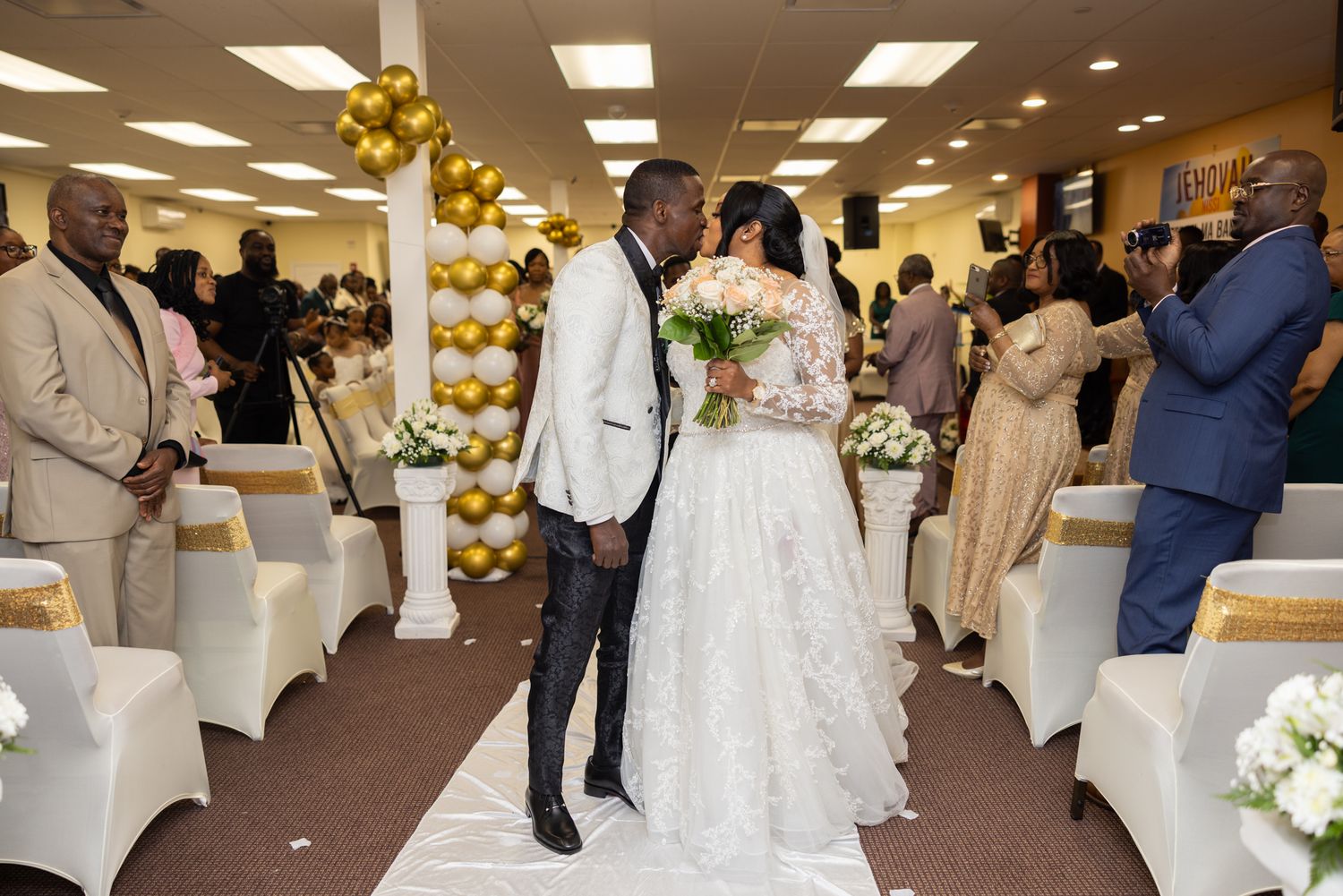 Wedding celebration in a reception hall decorated with gold balloons and white chair covers.