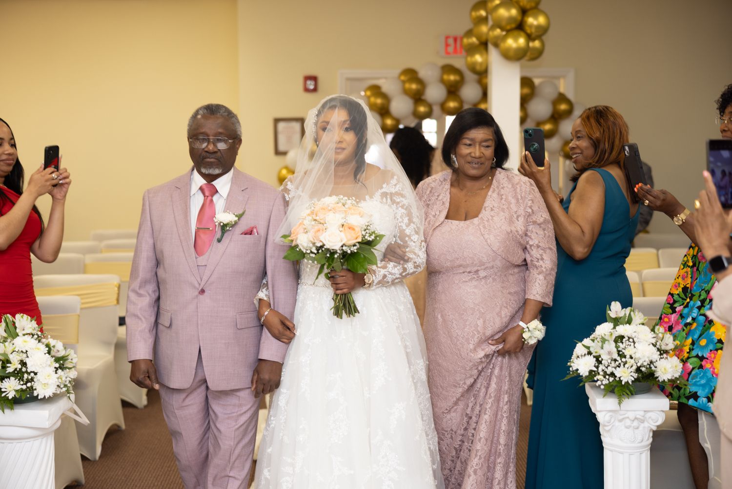 A wedding procession walks down the aisle with guests taking photos in a venue decorated with gold balloons.