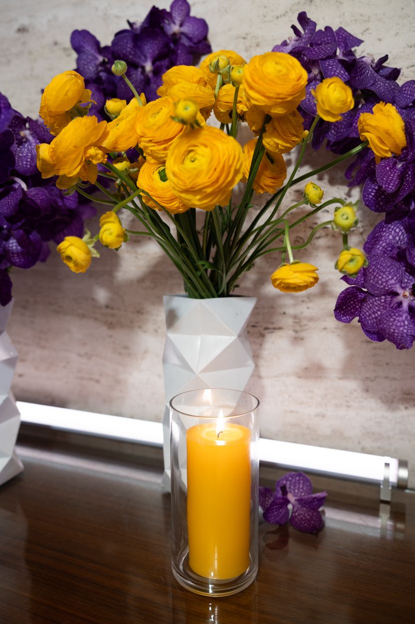 Yellow ranunculus and purple orchids frame a glowing candle in a glass holder on a wooden surface.