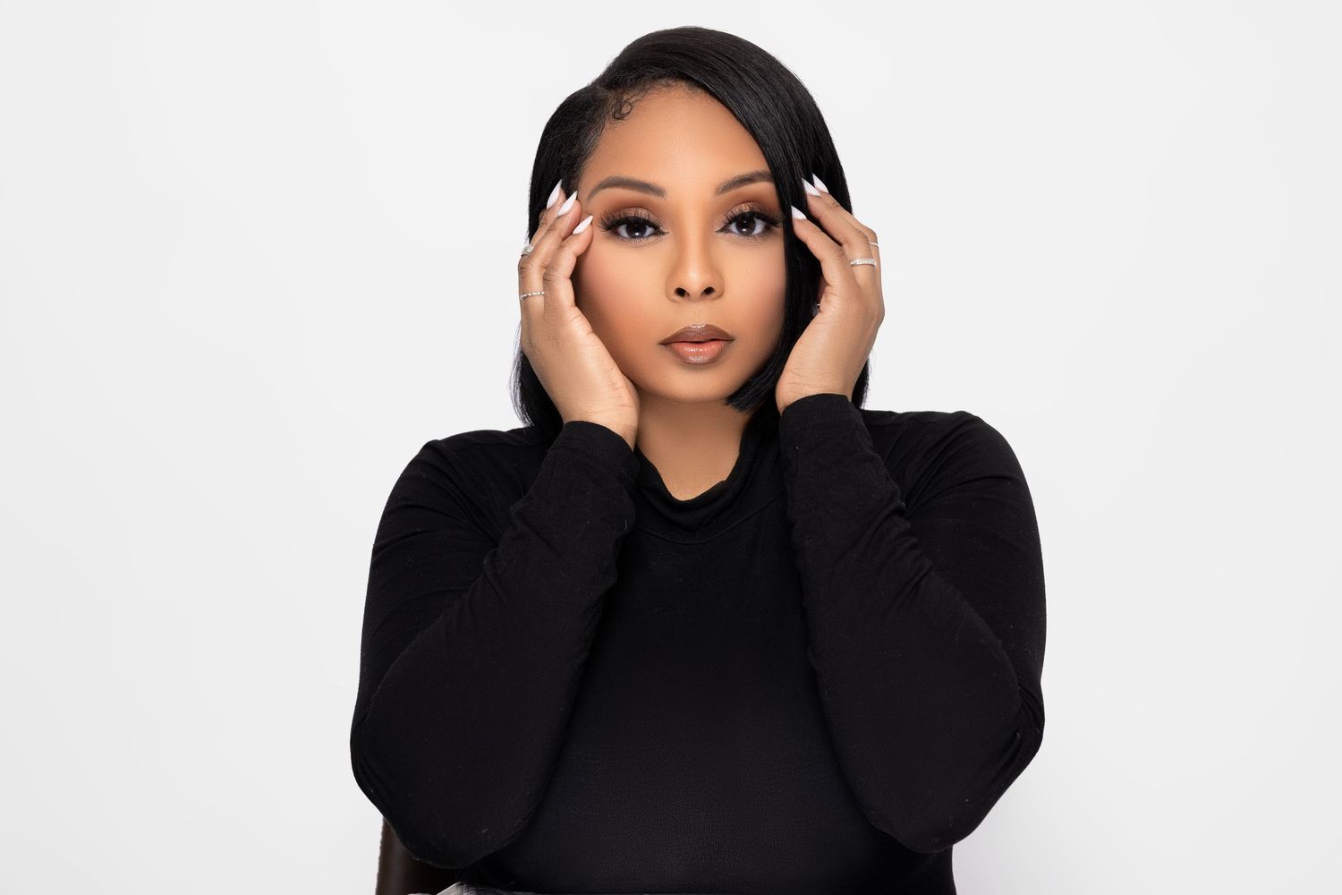 A person wearing a black turtleneck sweater poses with hands near their face against a white background.