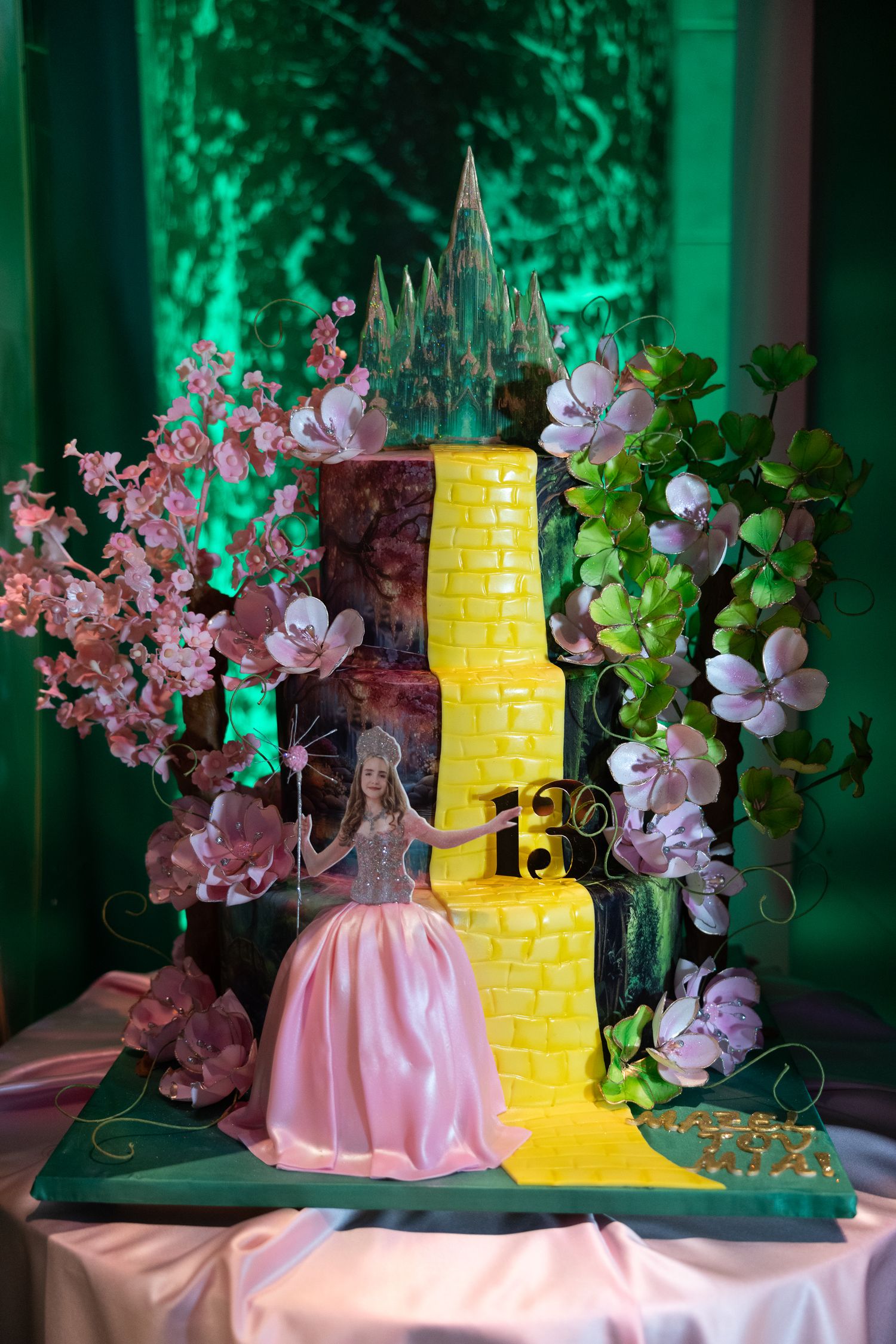 A pink and yellow wedding cake decorated with cherry blossoms and green foliage sits on a display table with emerald lighting.