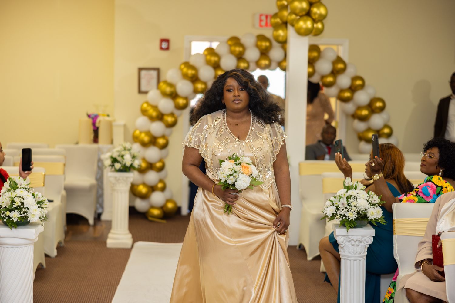 Wedding reception decorated with gold balloons and white floral arrangements in an indoor venue.