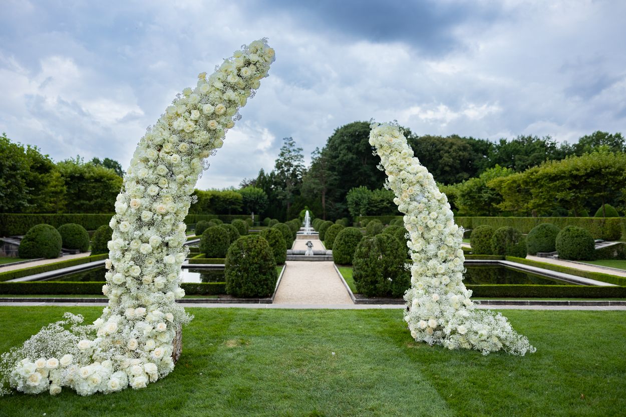 White floral wedding arch and ceremony chairs set up on manicured lawn with trees in background.