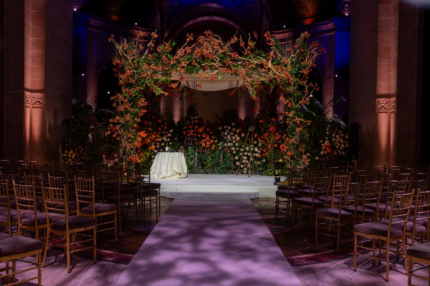 Wedding venue interior with purple lighting, red floral arrangements, and elegant gold chairs set up for ceremony.
