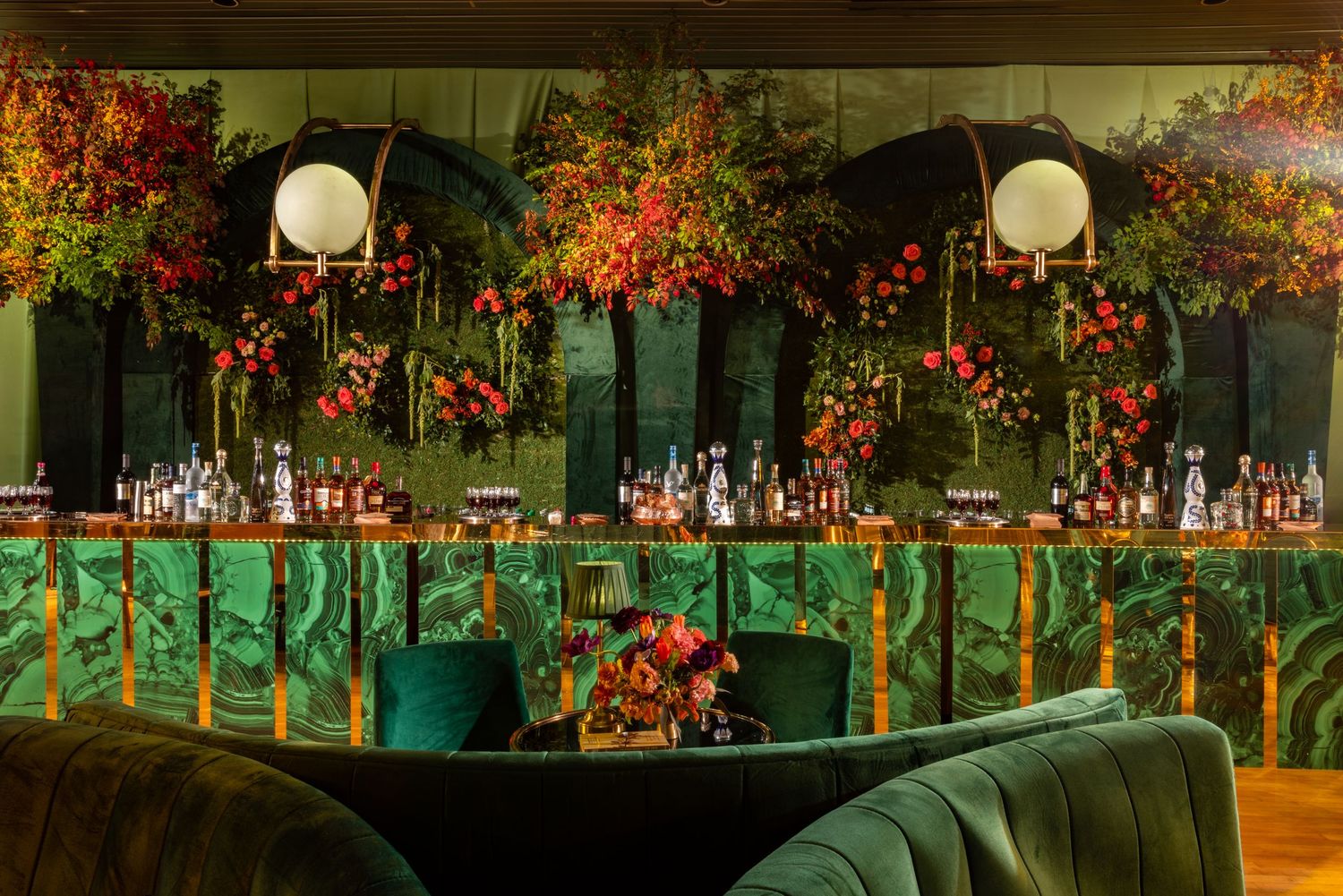 Luxurious bar setup with emerald lighting, velvet seating, and ornate autumn floral arrangements featuring glowing orb lights.