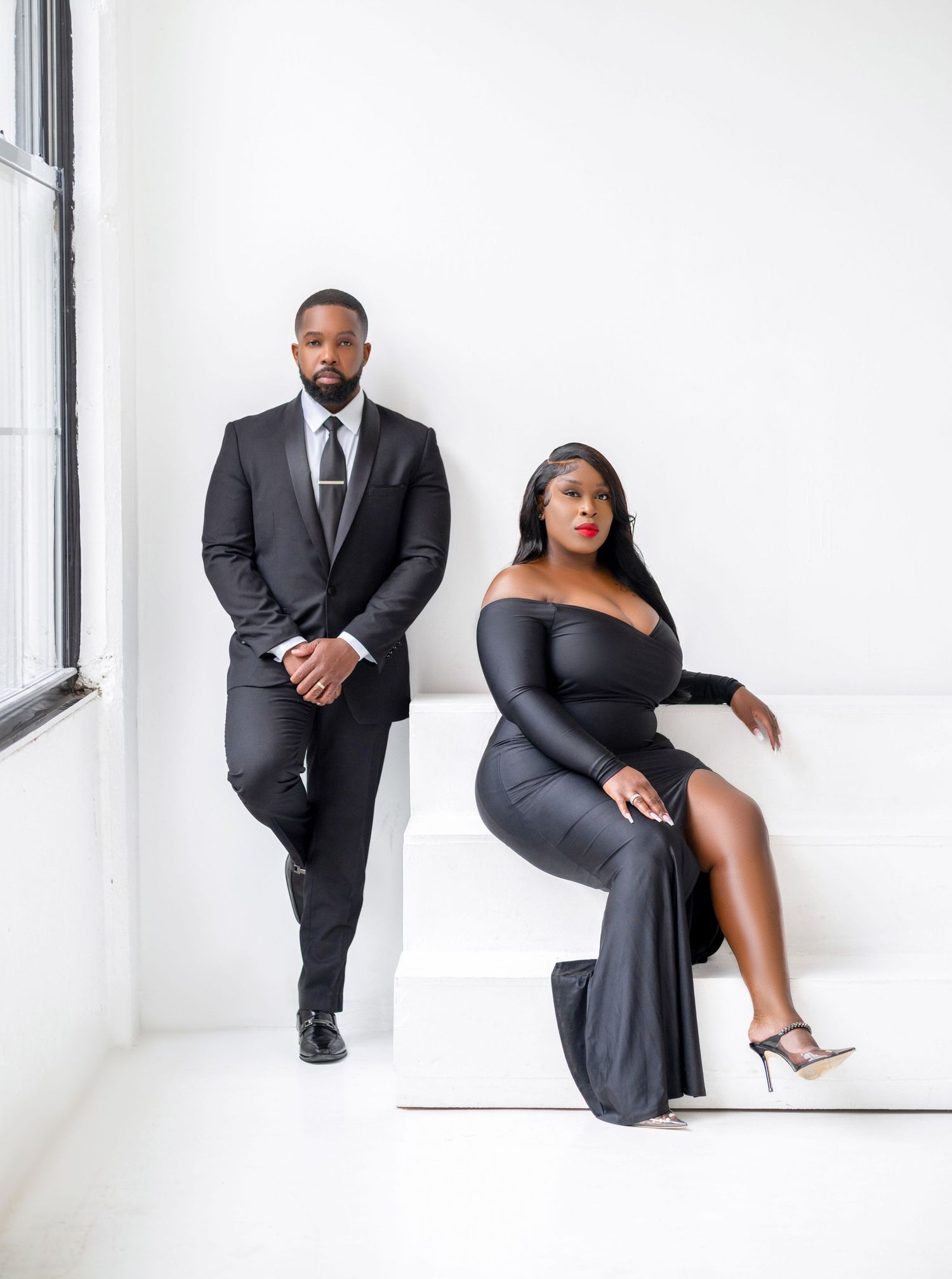 Elegant couple poses in formal black attire for a stylish studio photoshoot with white backdrop.