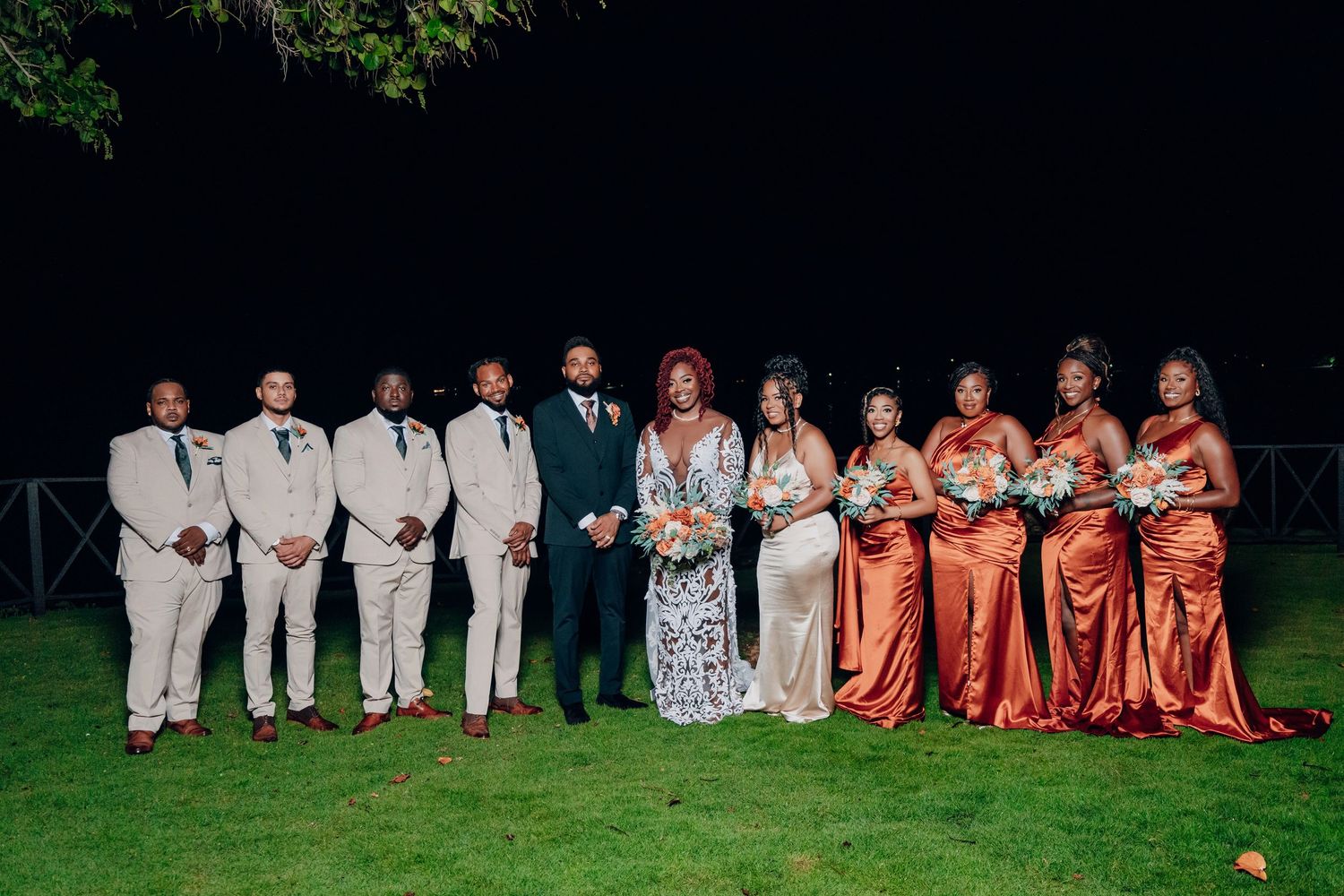 Wedding party members in white and coral dresses pose together on a grassy lawn at dusk.