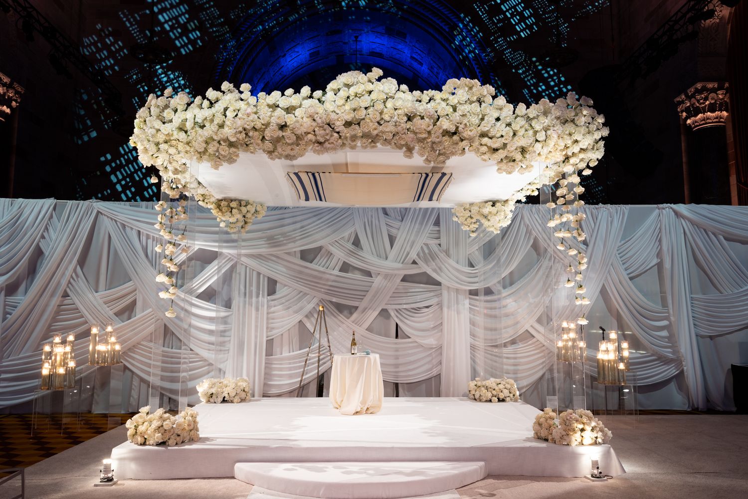 Elegant white wedding ceremony setup with ornate chandeliers and floral arch decorations against a draped backdrop.