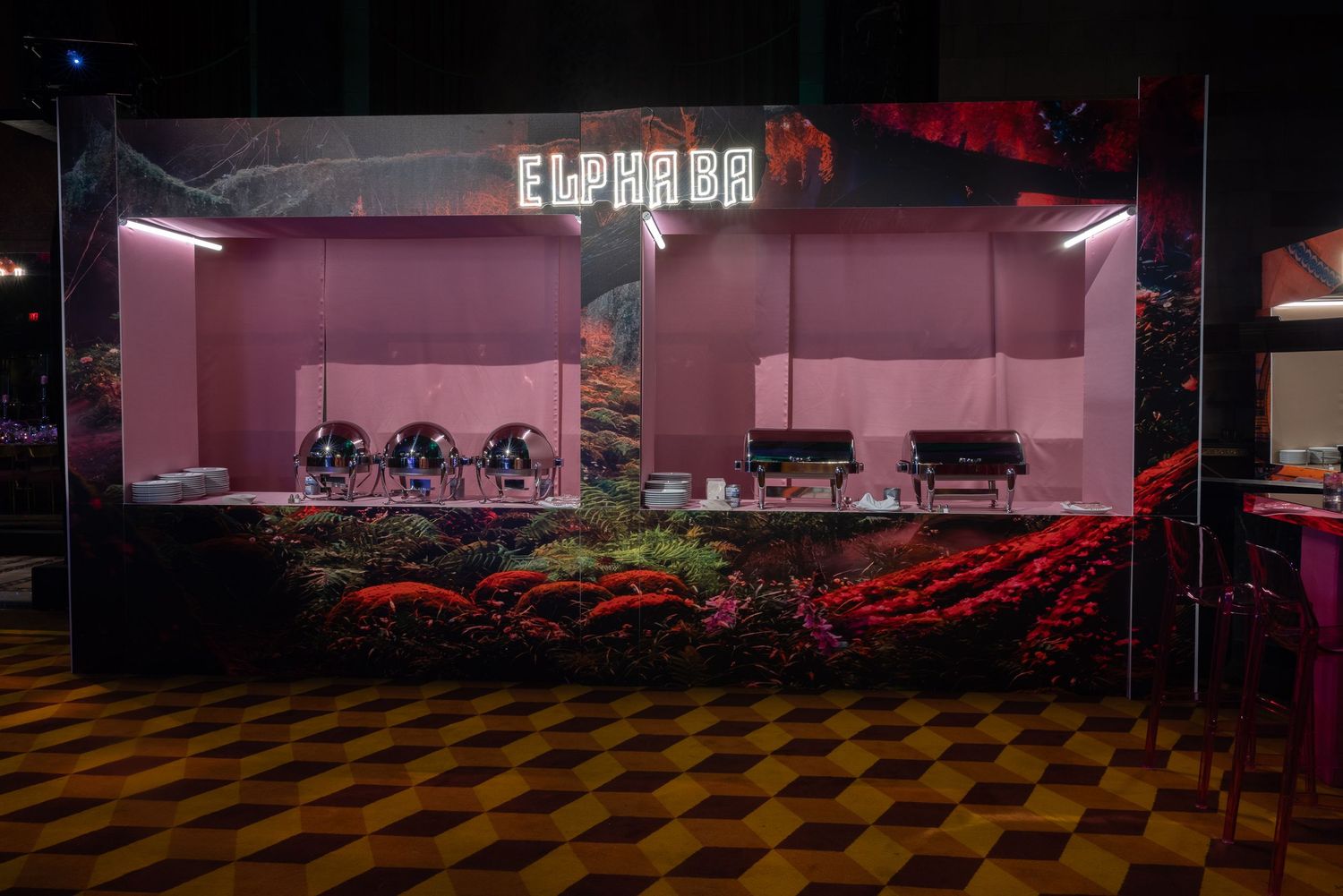 An empty event venue with pink lighting and checkered flooring features rows of chairs arranged in a banquet style setup.