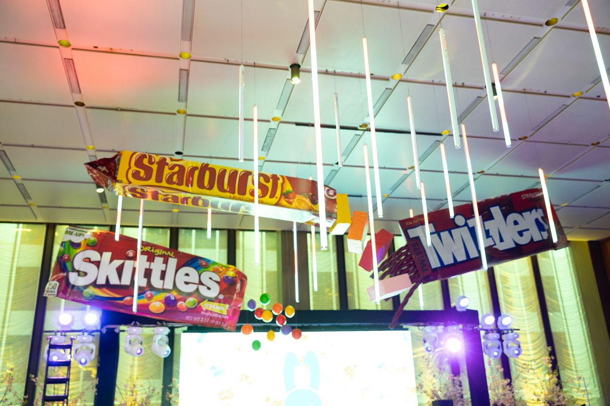 Colorful neon signs illuminate a retail storefront with candy and snack advertisements displayed in the window.