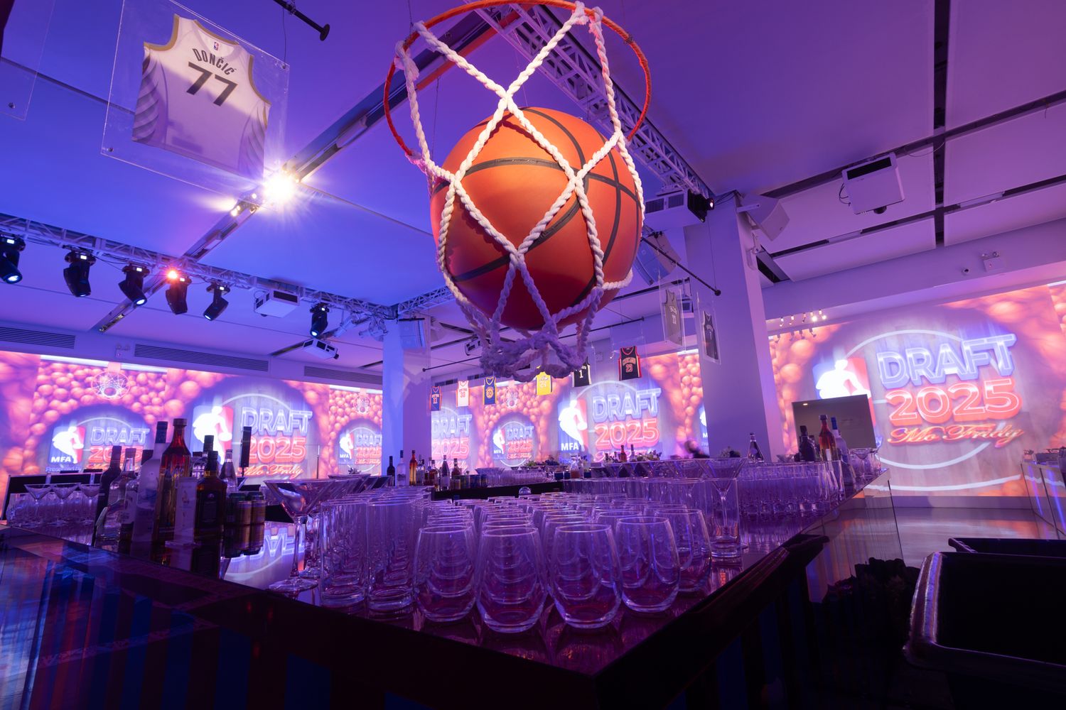 Purple and pink uplighting illuminates a large event space with white chairs and decorative ceiling installations.