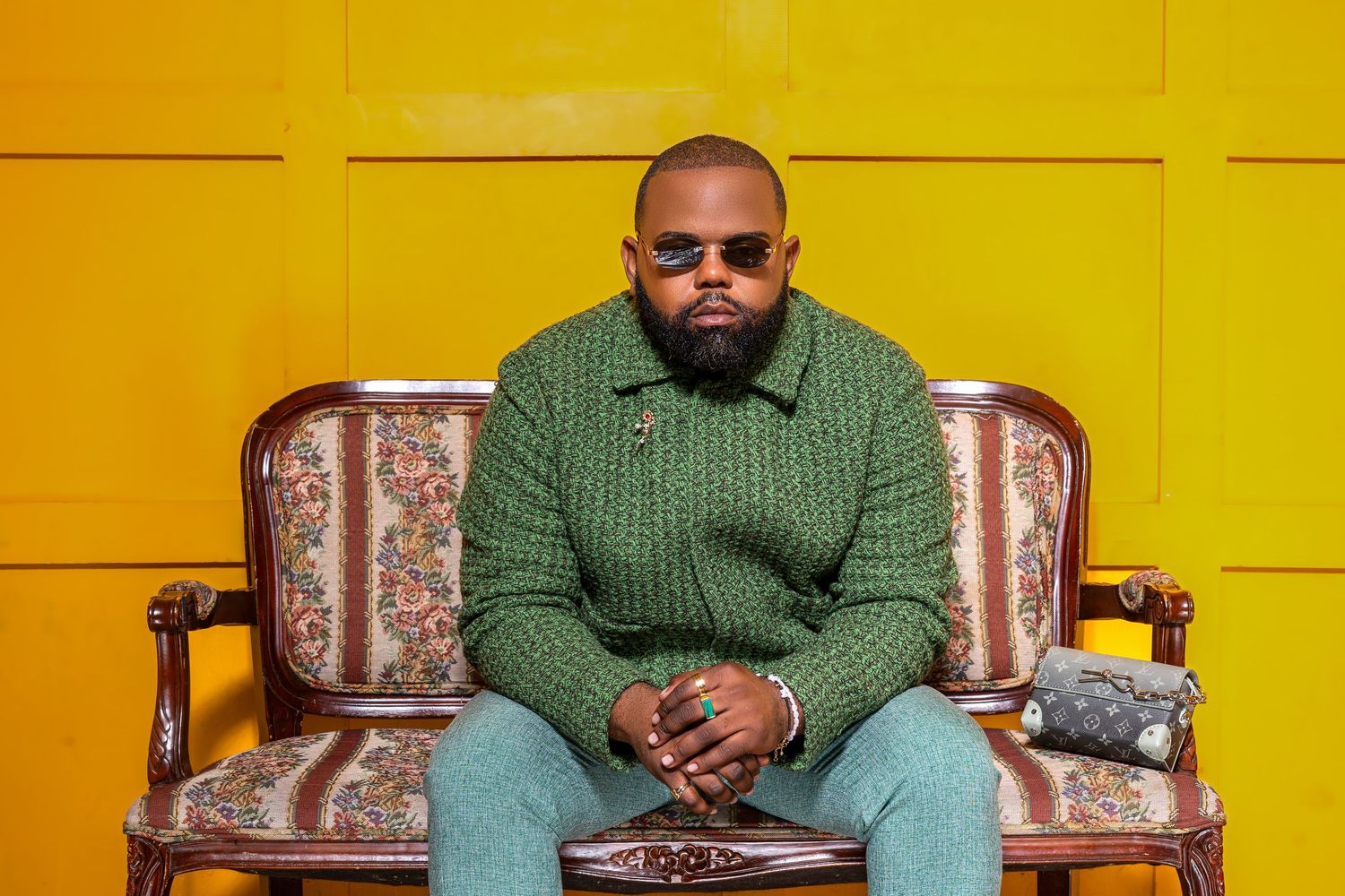 Person in green sweater sits on ornate chair against vibrant yellow wall in studio setting.