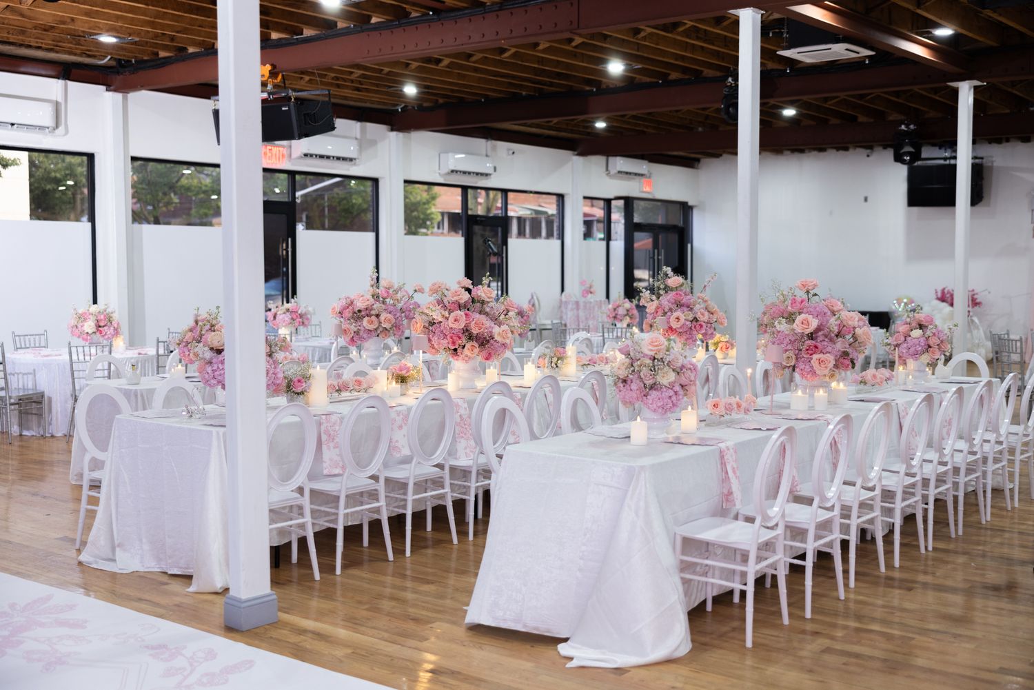 Elegant wedding reception venue with long white tables, pink floral centerpieces and white chiavari chairs on hardwood floors.
