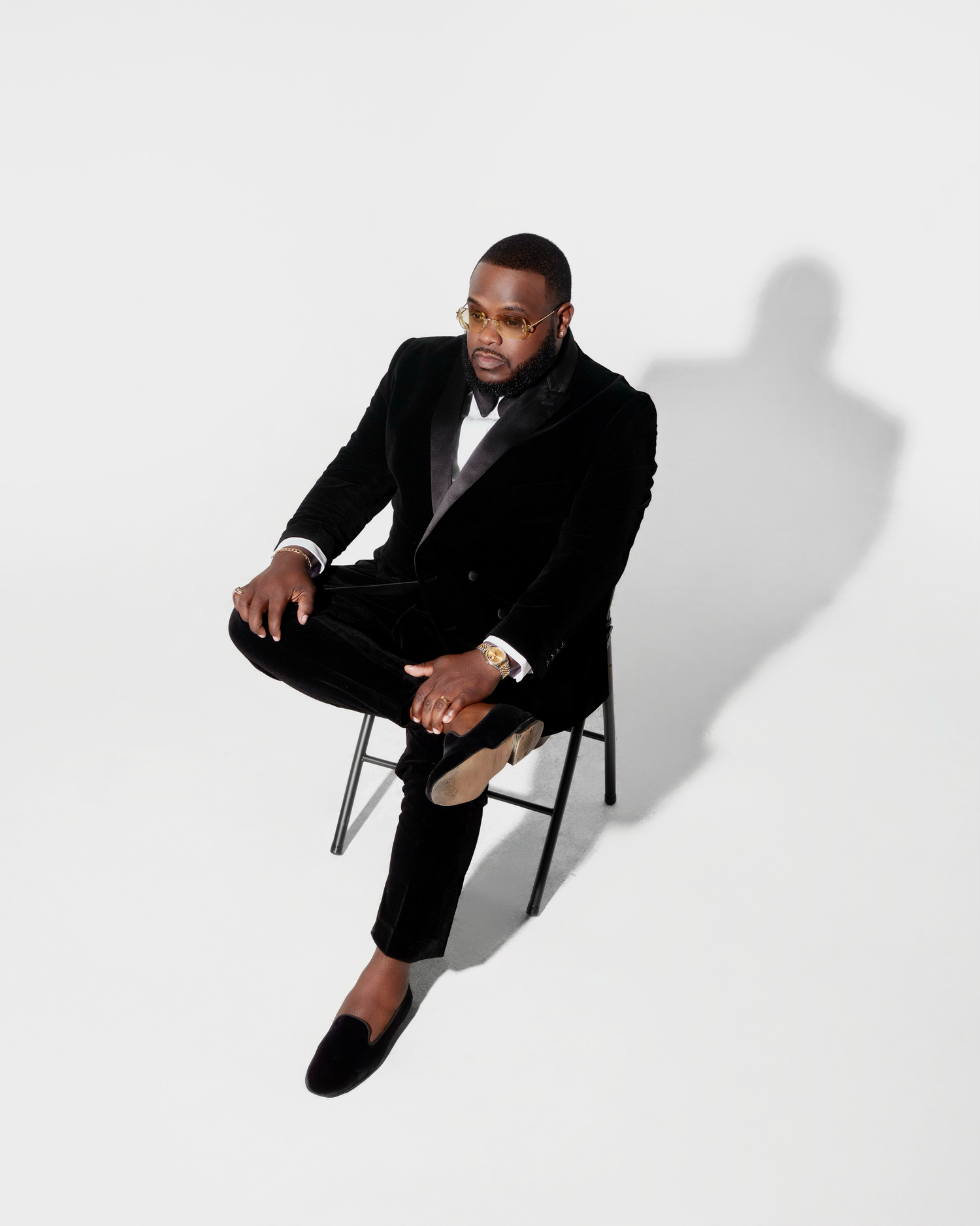A person in a black tuxedo sits on a chair against a white background casting a dramatic shadow.