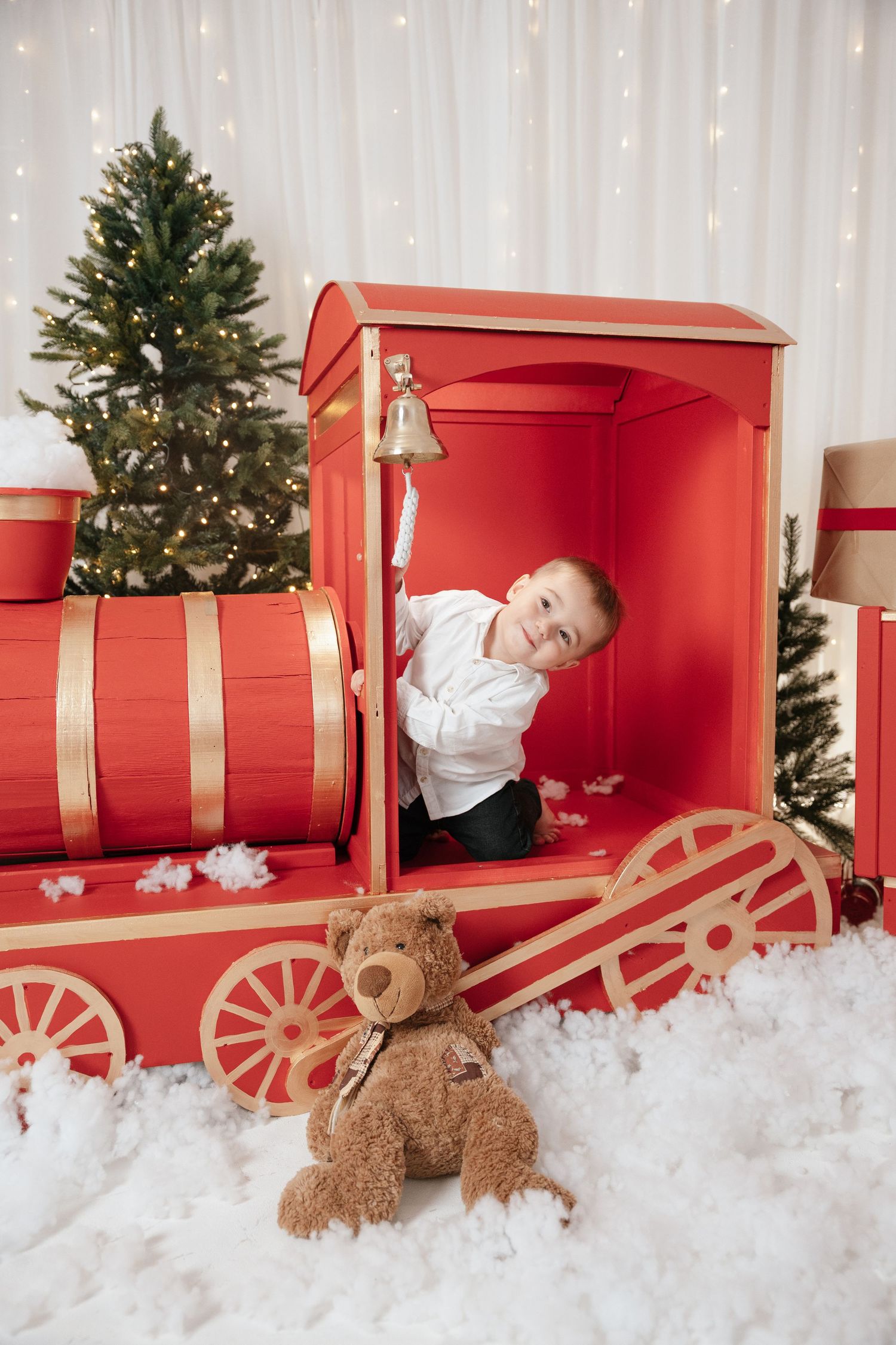Boy smiling in red toy Christmas train with teddy bear, snow, and decorated trees in festive holiday photo setting.