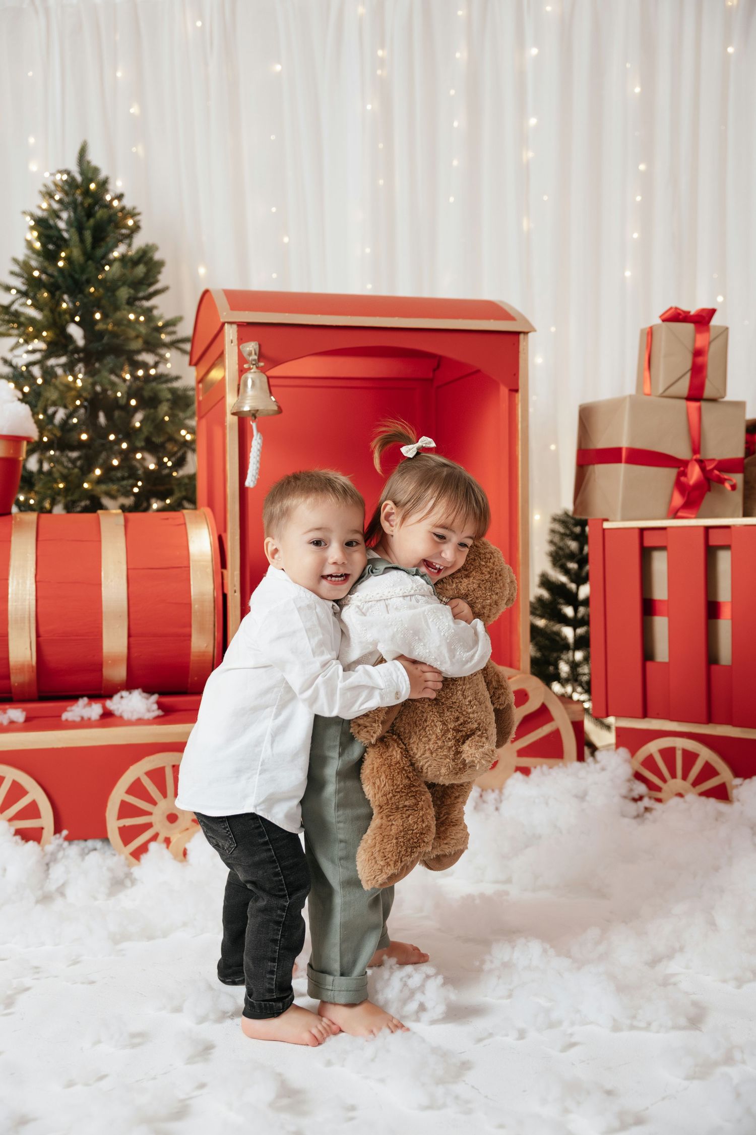 Two young children hug while holding a teddy bear in a festive Christmas setting with gifts and a red toy train.