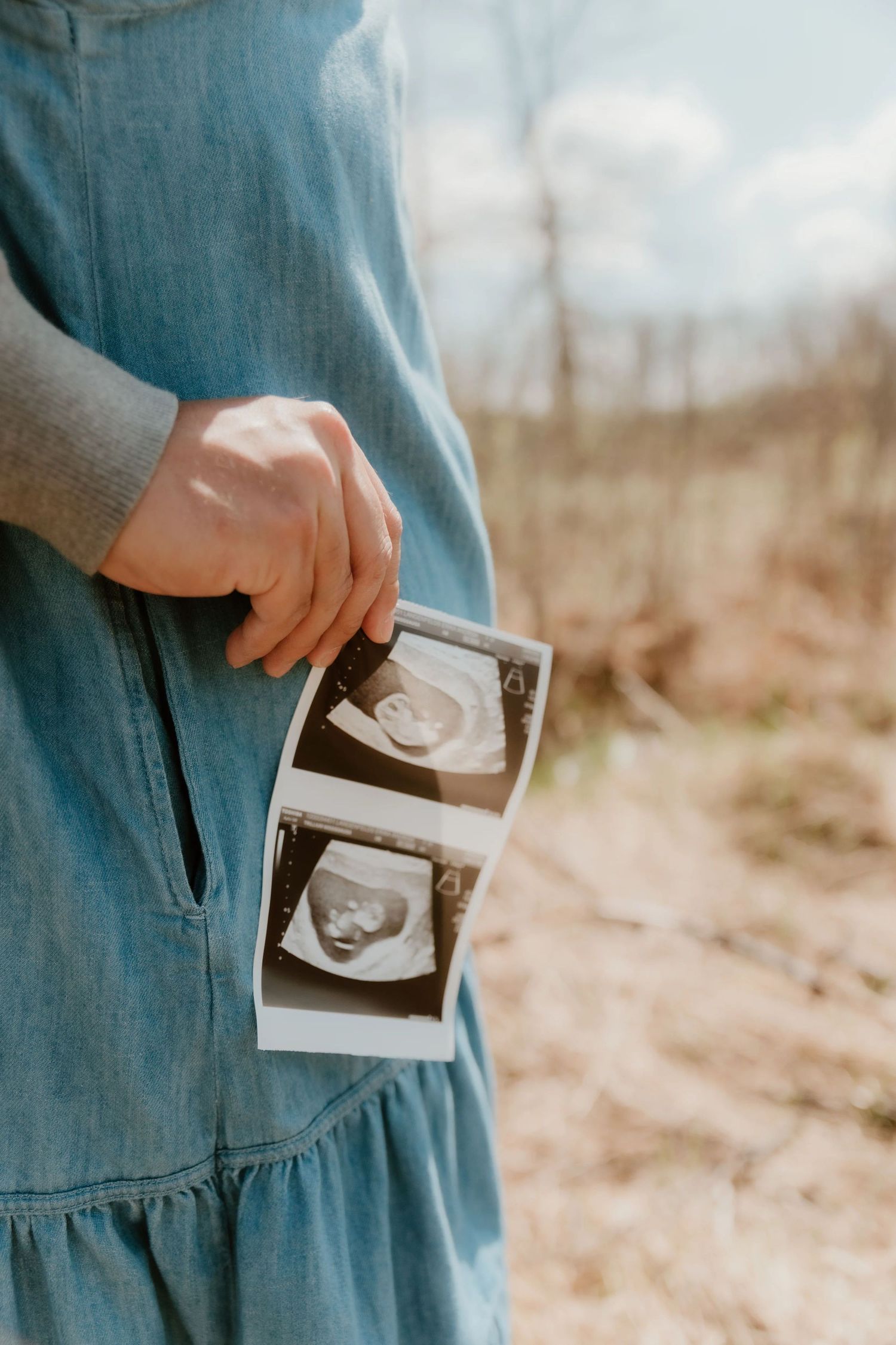 Someone in a blue dress holds ultrasound sonogram photos while standing outdoors on a sunny day.