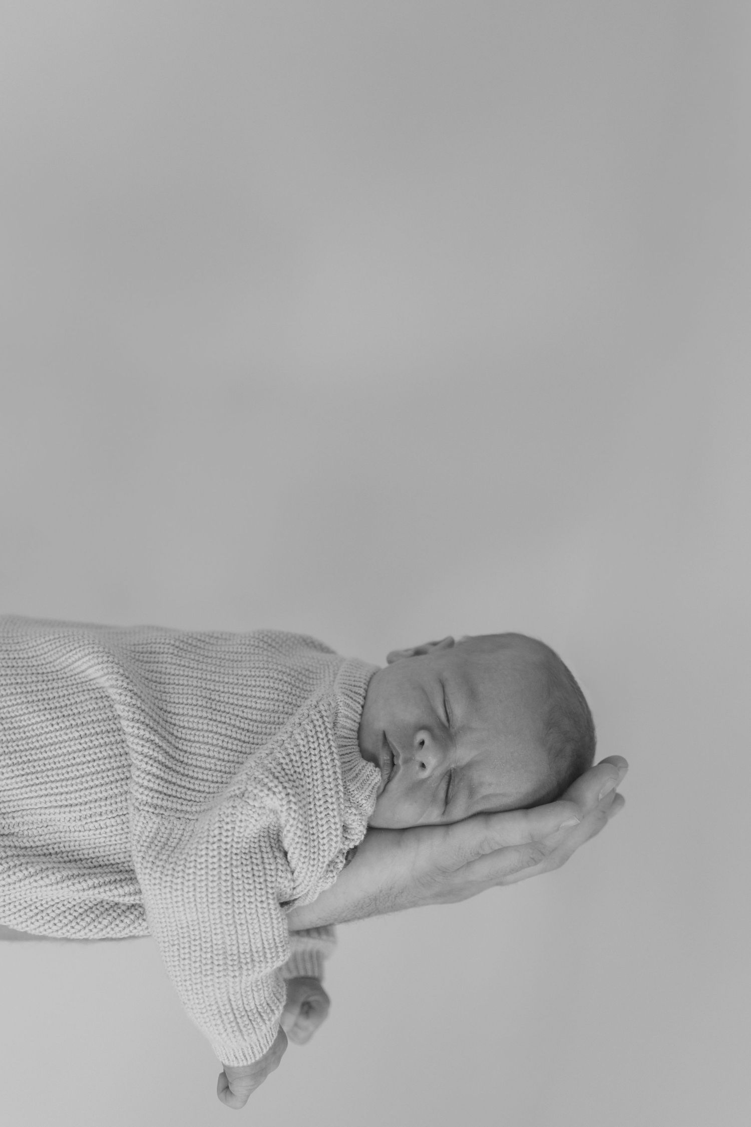 Black and white newborn photo of a sleeping baby in a knit sweater resting peacefully on an adult's open hand.