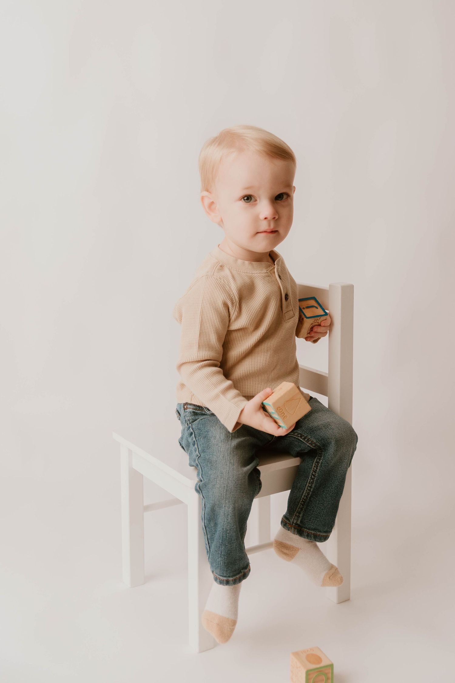 Small child wearing a beige sweater and jeans sits on a white chair against a light background in a minimalist setting.