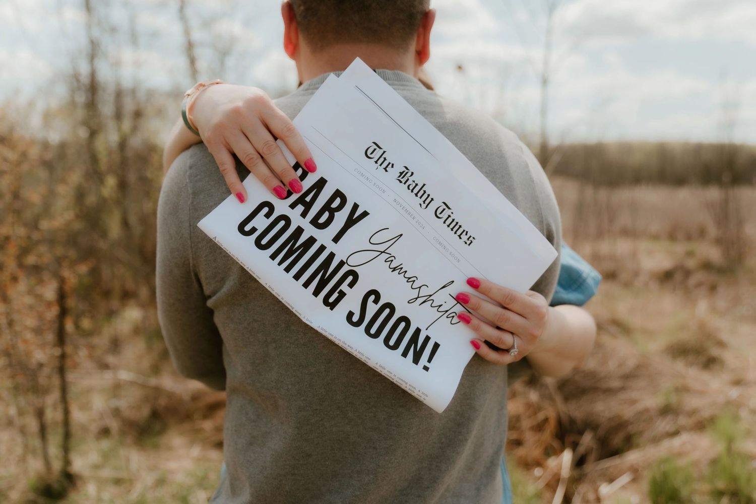 Someone holds a 'Baby Coming Soon' announcement sign in a natural outdoor setting against a blurred background.
