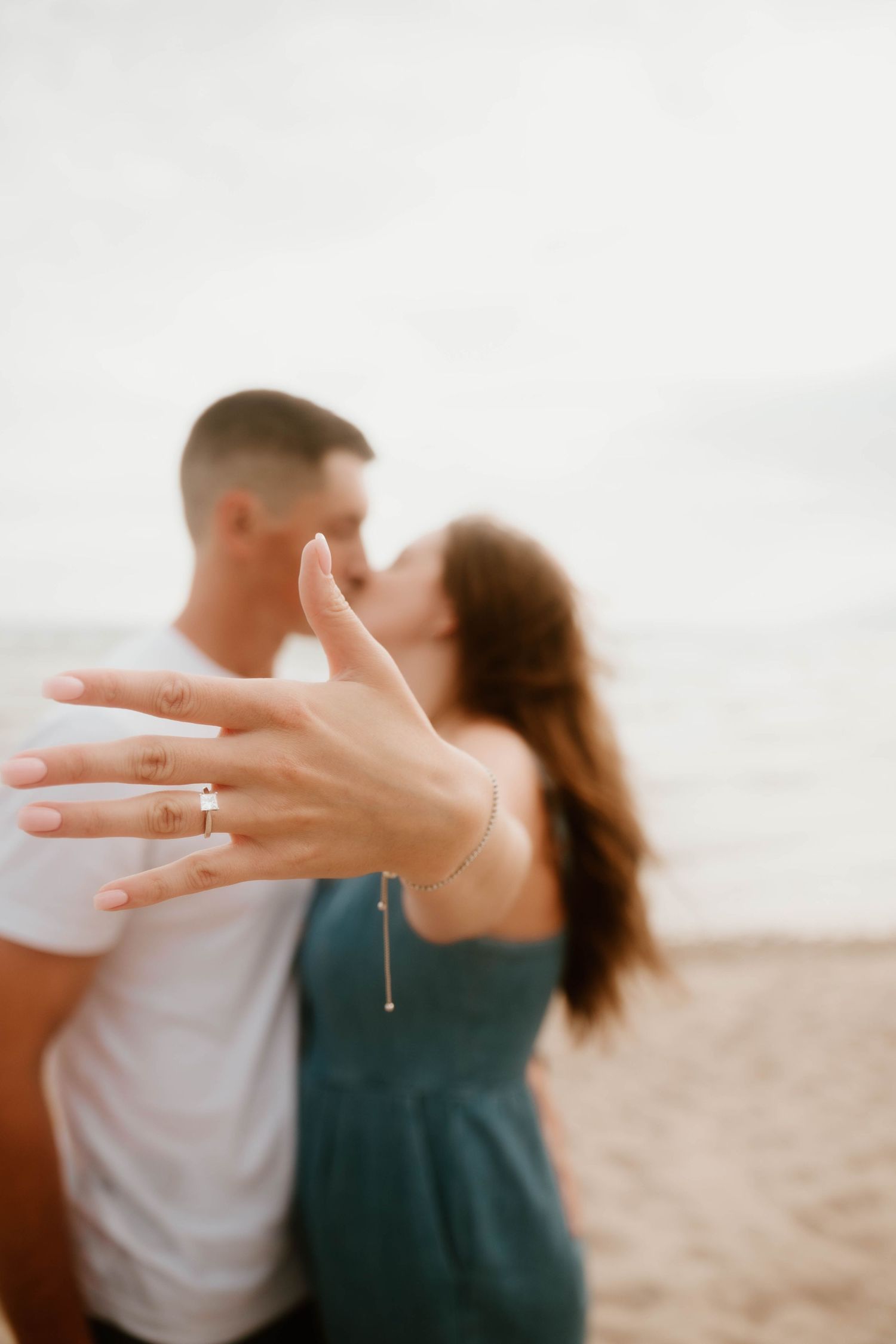 Engagement ring displayed on hand with couple embracing in soft focus background.