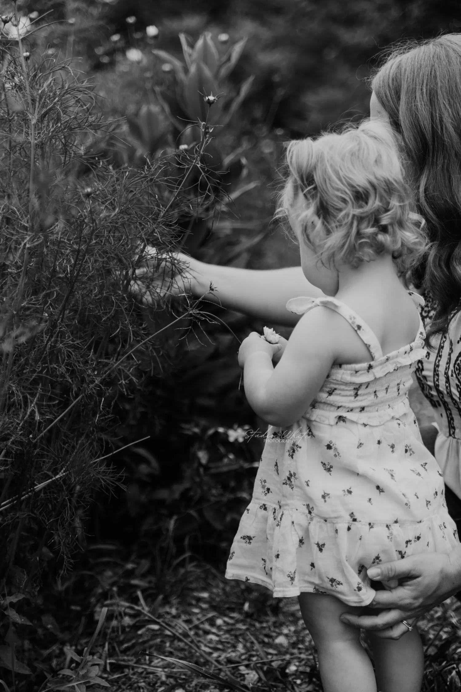 A child in a patterned dress reaches out to explore plants in a garden, captured in black and white photography.