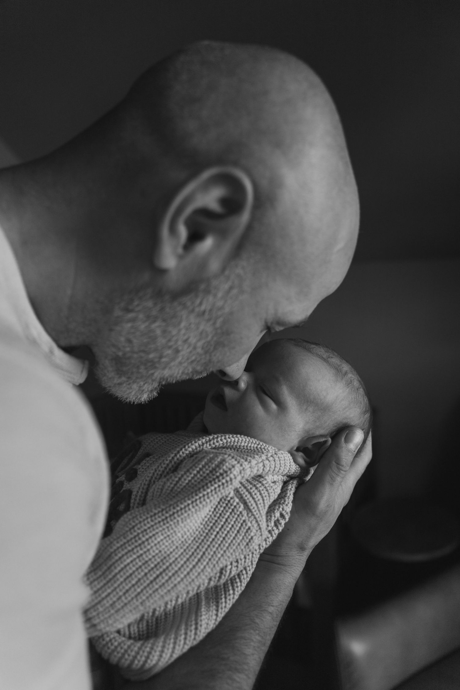 Black and white photo of a bald, bearded father tenderly kissing the forehead of his sleeping newborn baby wrapped in a knit blanket.