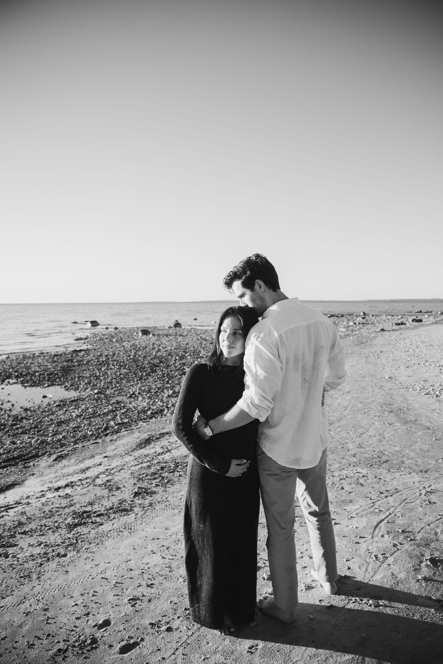 Black and white photo of a couple embracing on a rocky beach, the man in a light shirt holding the woman in a dark dress by the sea.