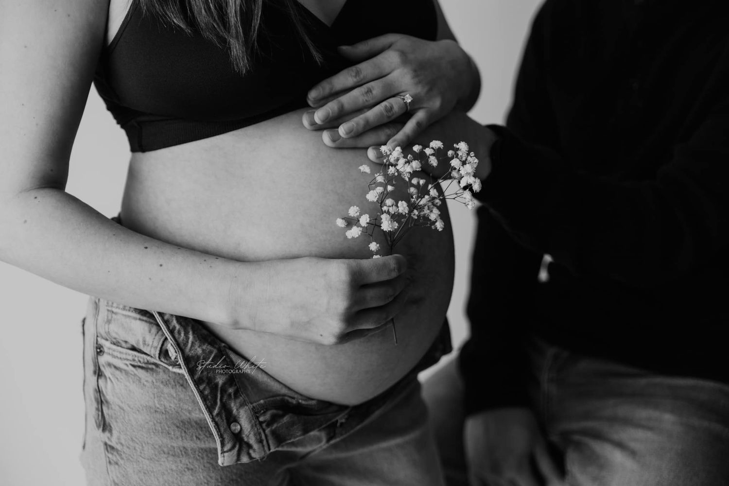 Close-up of womens pregnant belly wearing a sports bra and with hands holding babys breathe flowers, husband hands on belly.