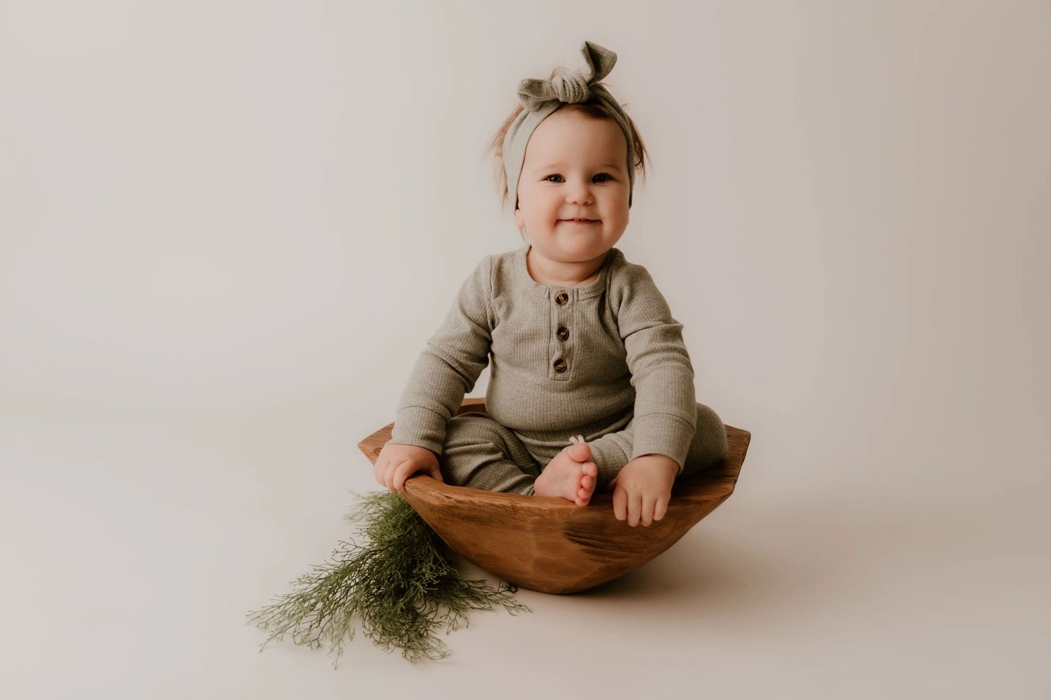 Young child in gray outfit sits in wooden bowl with greenery.