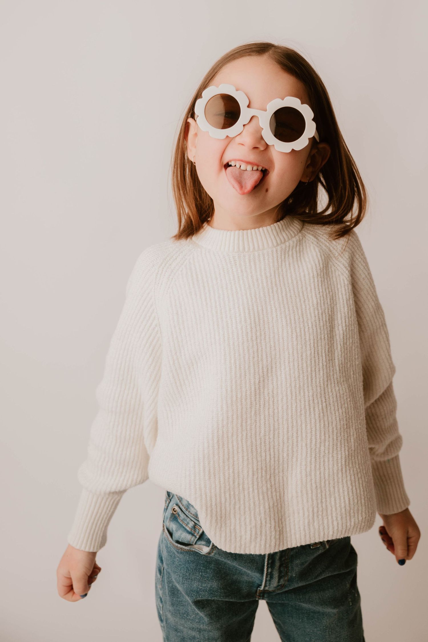 Child wearing white knit sweater, jeans and round sunglasses poses playfully against light background.