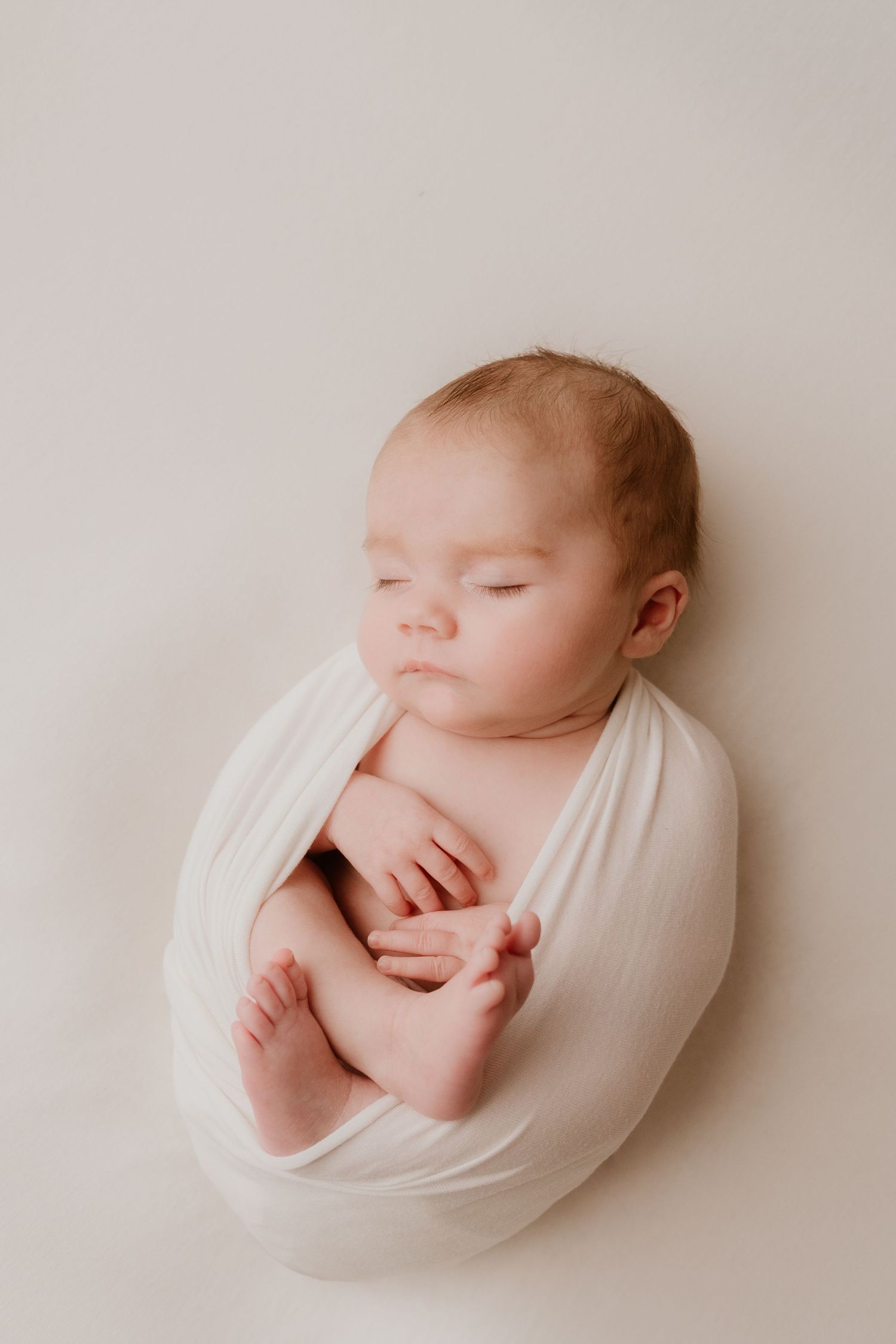 Peaceful newborn baby swaddled in white wrap sleeping on cream background in professional studio photo.