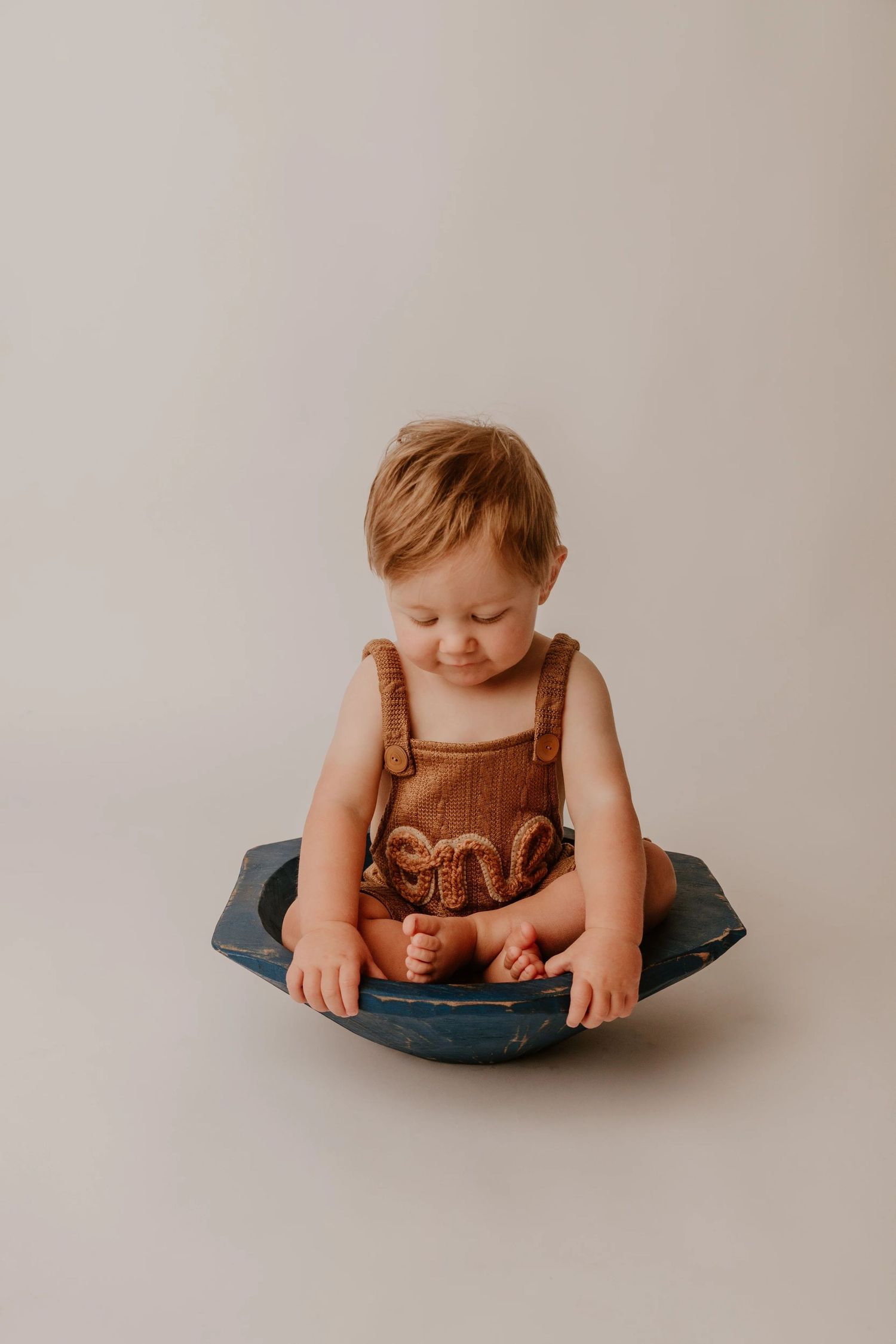 Small child wearing brown overalls sits in a round blue bowl against a neutral background.