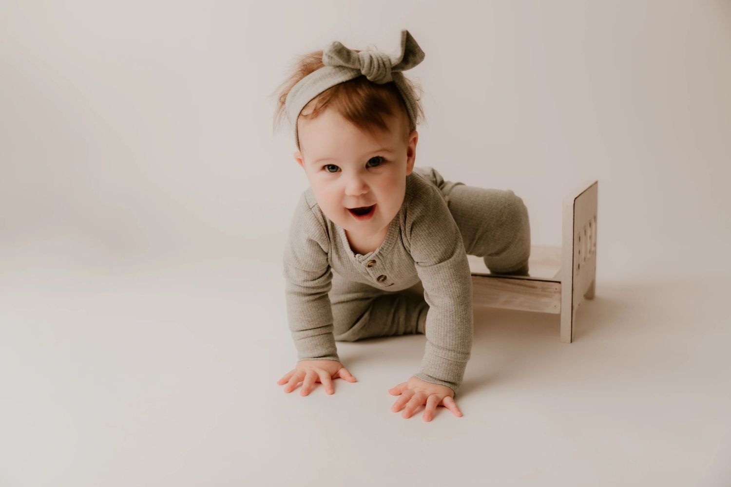A baby in a neutral-colored onesie crawls and plays on a light beige floor in a minimalist setting.