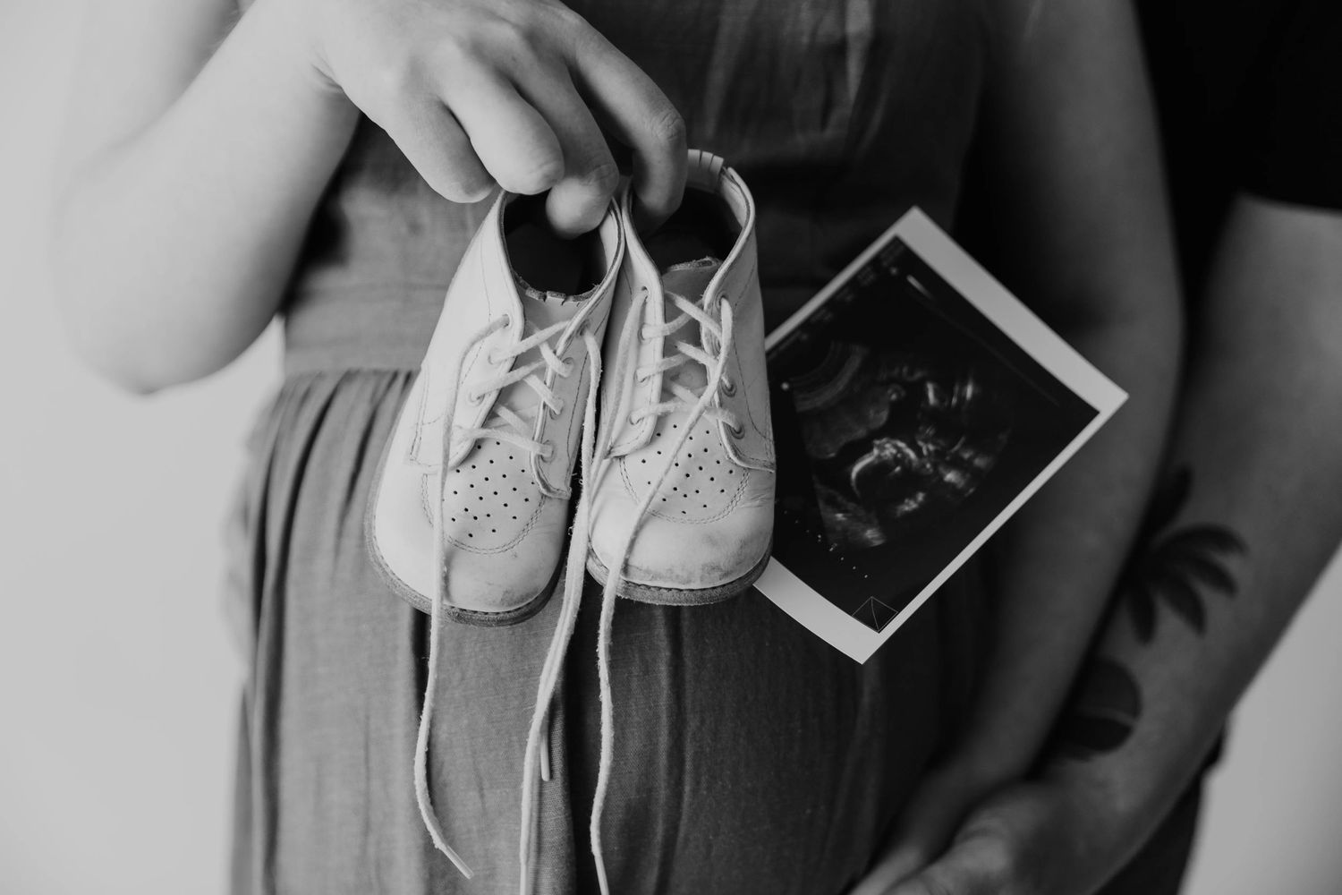 A close-up of baby white sneakers with laces being tied against women's pregnant belly, behind it is an ultrasound photo.
