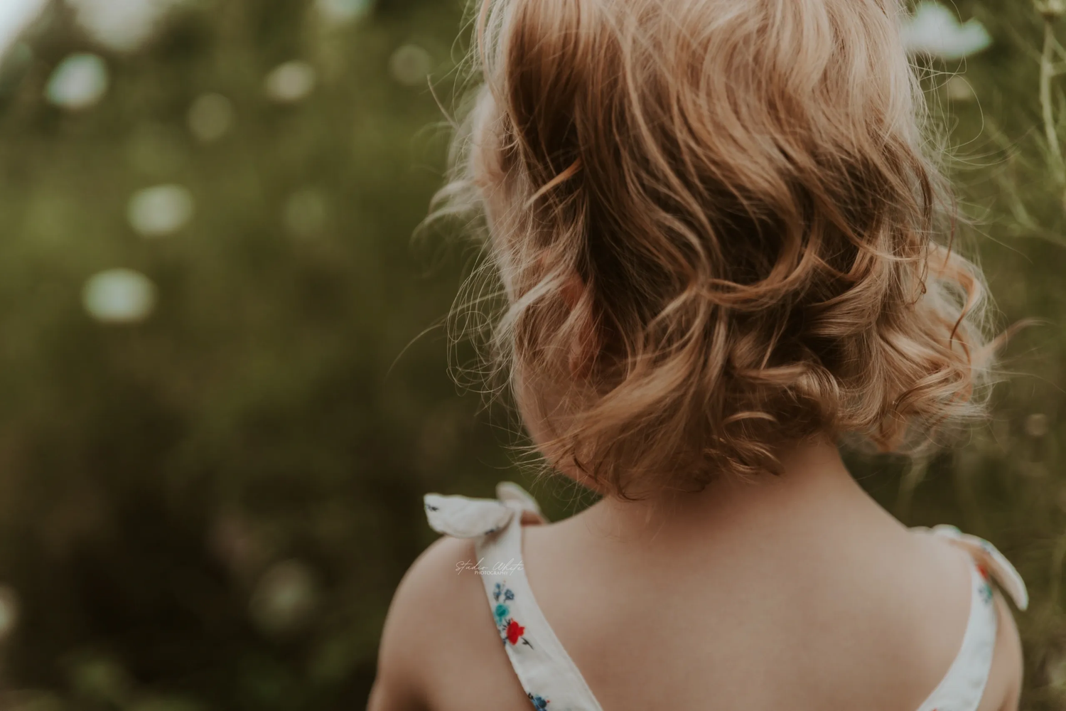 A toddler with shoulder-length wavy brown hair is shown from behind against a blurred natural background.